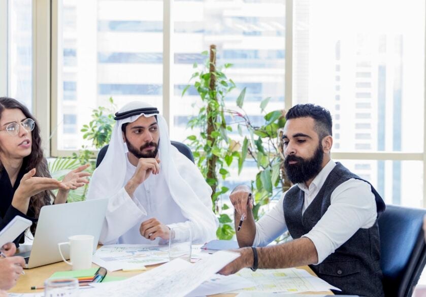 Five business professionals sat round a meeting table with one of the professionals wearing traditional Islamic clothing.