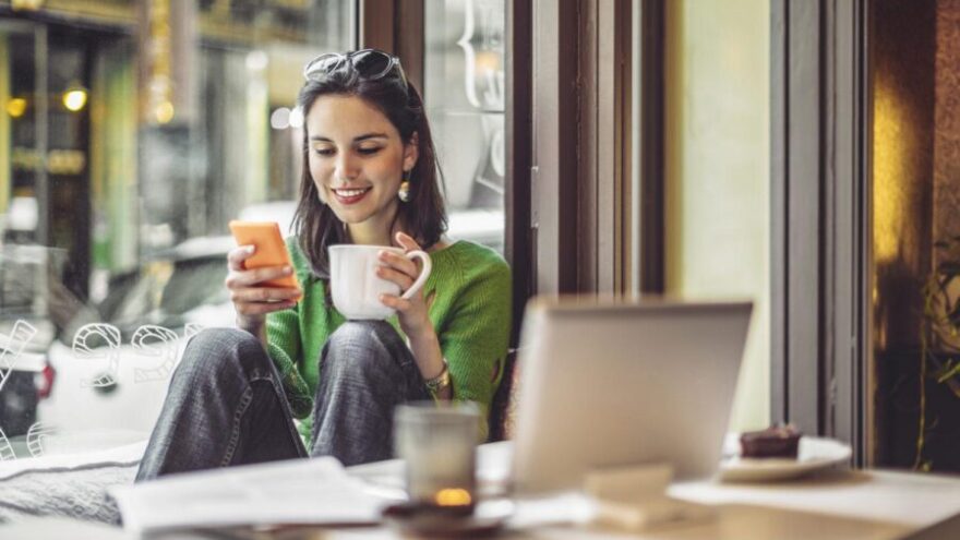 Woman working from a cafe with a coffee and phone in hand, taking advantage of the workplace's flexible working policy.