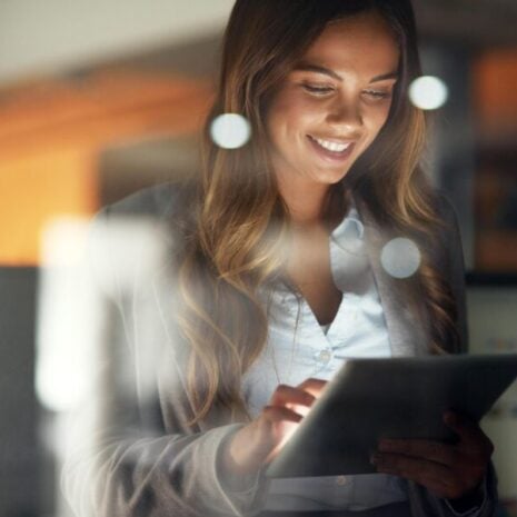 An EA holding an iPad in a darkened office while working. She's smiling and happy with her job.