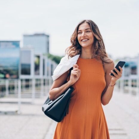A female chief of staff walks with her phone in hand on a sunny day in the City. She's carrying a black leather handbag.
