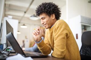 Side view if happy young businessman sitting at his desk and working on laptop computer. Egyptian male professional looking at his laptop and smiling in office.