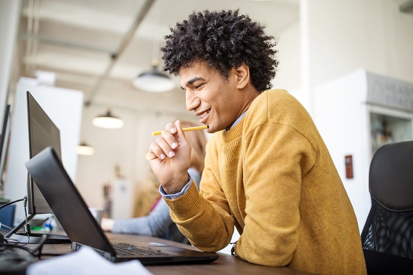 Side view if happy young businessman sitting at his desk and working on laptop computer. Egyptian male professional looking at his laptop and smiling in office.
