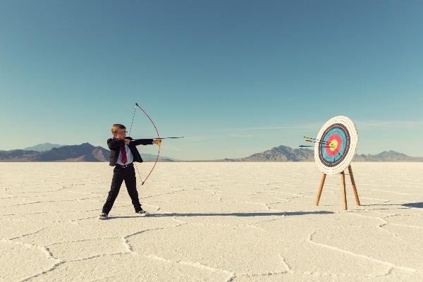 A little boy using a bow and arrow to shoot a target in the middle of a hot and sunny desert.