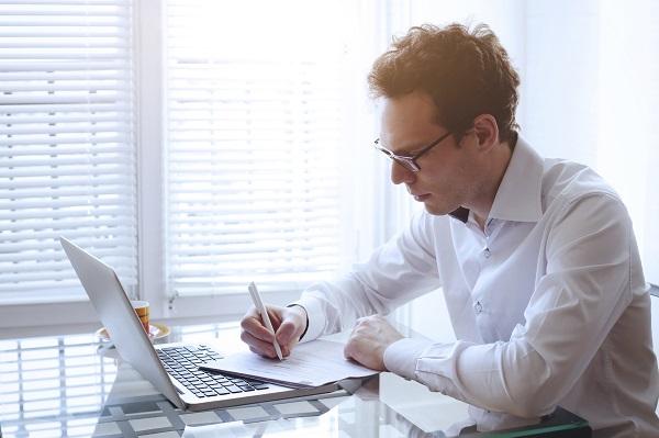 A man with tousled hair and glasses using a laptop and taking notes on a desk, searching for a job in a tough market.