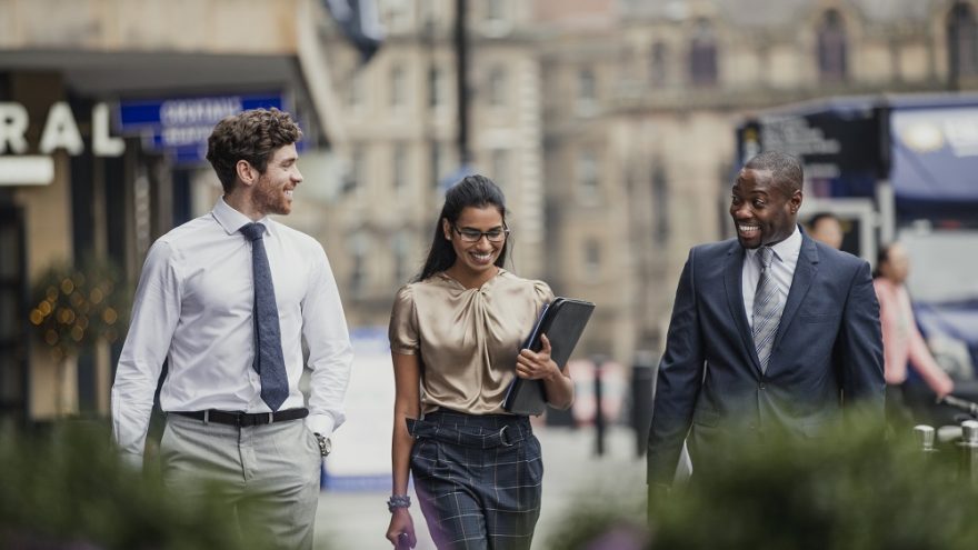 Three smartly dressed business people smiling and talking on city street.