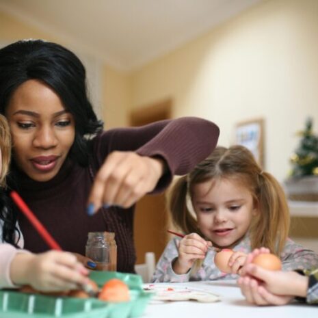 A nanny plays with three children, two girls and a boy, helping them paint eggs during an Easter celebration.