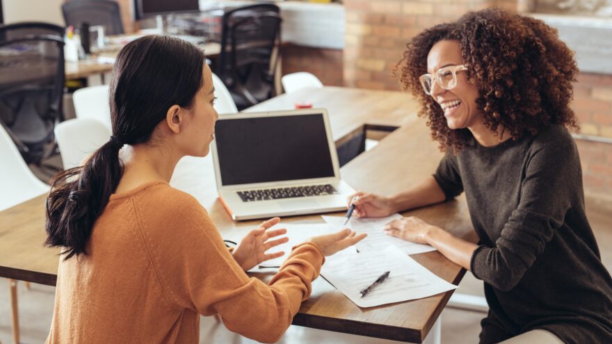 A HR professional onboarding a graduate in an open plan, casual office setting, sitting at a desk with a laptop.