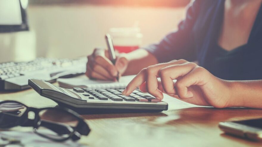 A woman deciphering her pension using a calculator, a computer, pen and paper while working in her office.