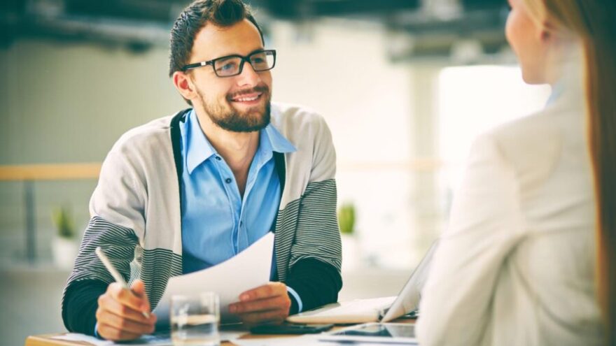 Man wearing glasses and smiling and looking at a colleague over a desk while conducting a performance review in an office.