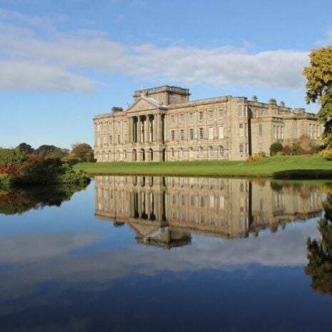 A large manor house where live-in couples work - there is a lake in the foreground and a few large trees surrounding it.