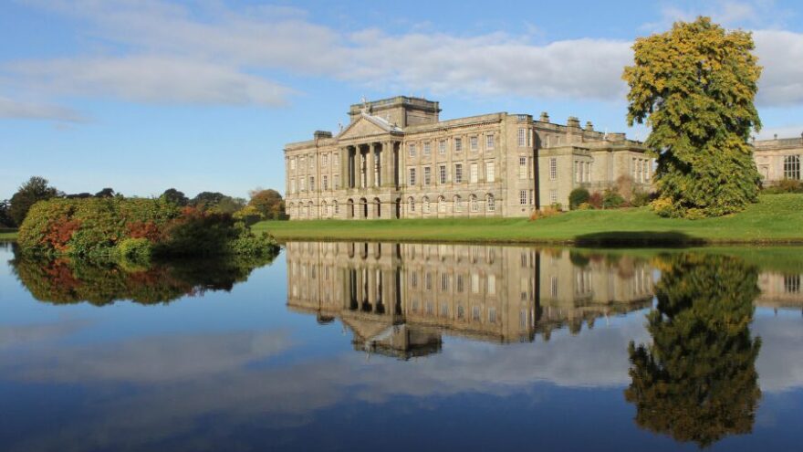 A large manor house where live-in couples work - there is a lake in the foreground and a few large trees surrounding it.