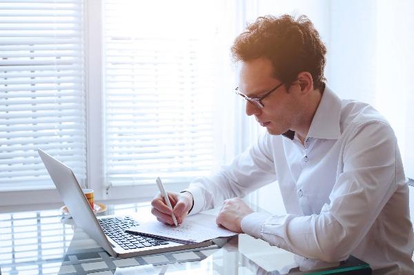A businessman in glasses sitting at his desk is writing his resume using a laptop computer and a piece of paper.
