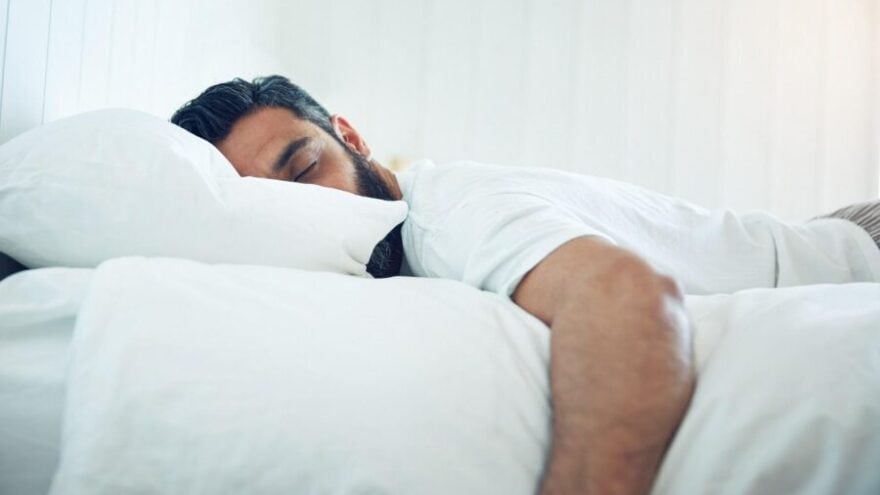 A man with dark hair and a beard in pyjamas sleeping on a white bed, bedsheet, duvet and pillow.