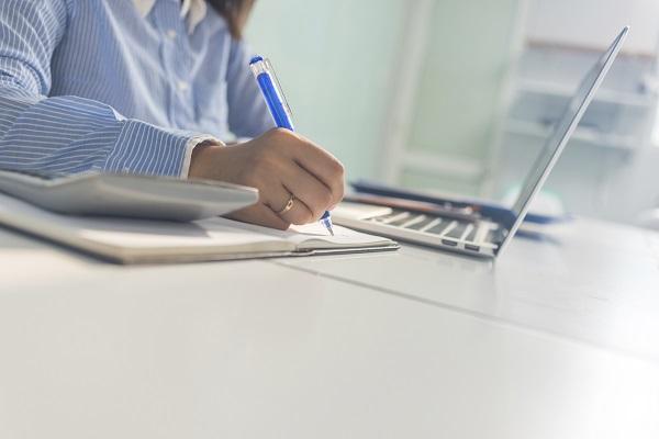 A close up of a woman using a laptop, pen and paper and a calculator on a desk in a sunny office.