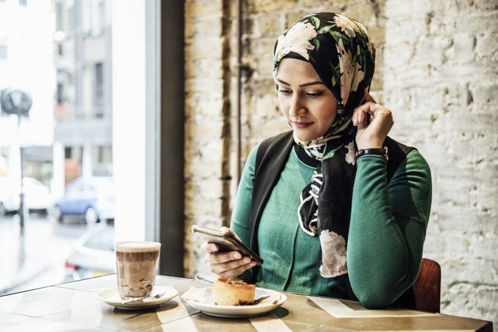 Woman in hijab texting on phone, hot drink and snack on table, leaning on elbow