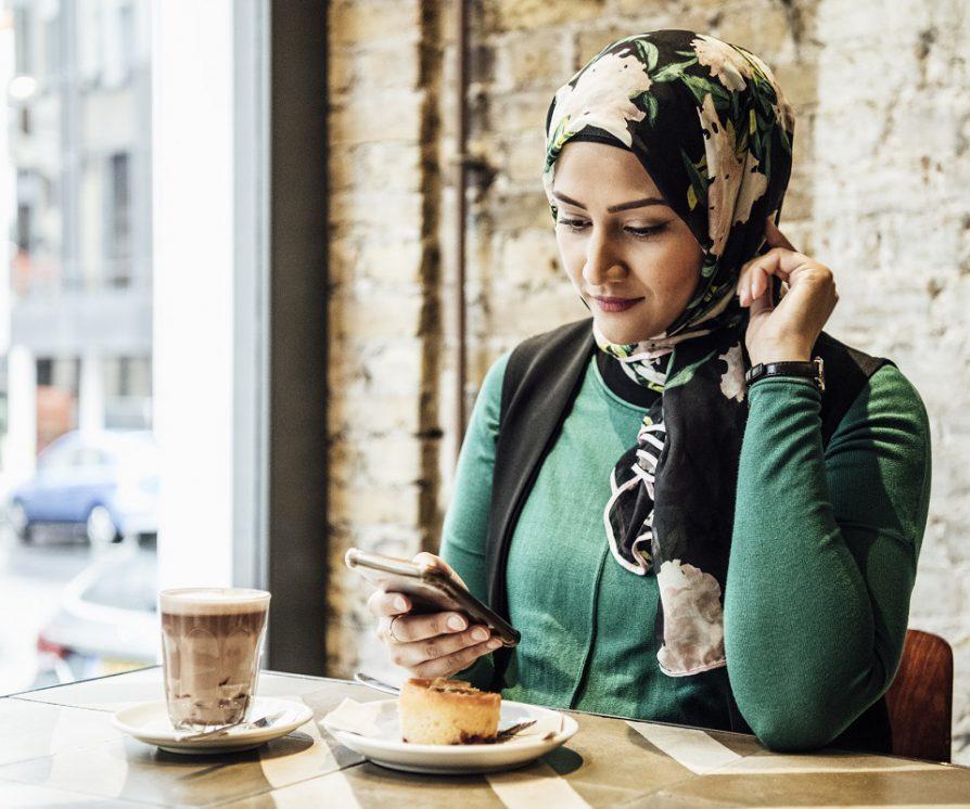A female human resources professional texting in a phone while working in a café, with a hot drink and snack on the table.