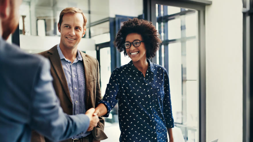 Three business people dressed in professional wear, two of them are shaking hands and smiling