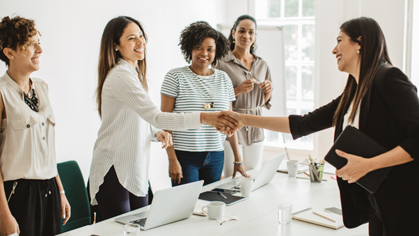 Women standing around table in meeting room, shaking hands and smiling.