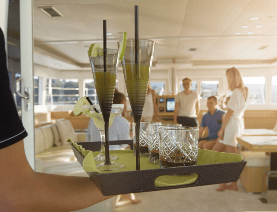 A temporary worker carrying a tray of drinks on a yacht towards a principal and their family.