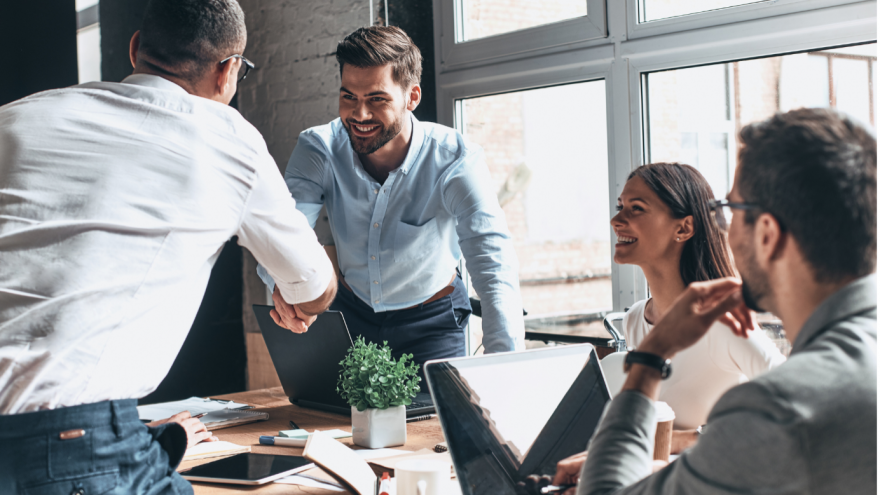 Four business professionals in a meeting room with two people stood, shaking hands